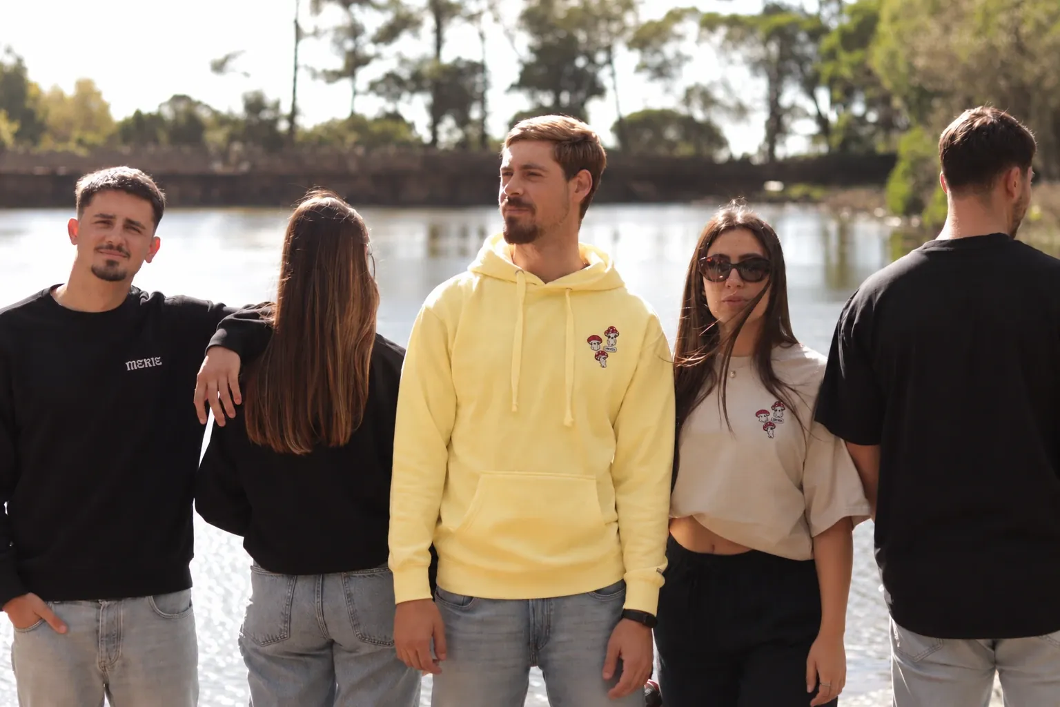 Amigos da Mekie junto a um lago, com hoodie amarelo, sweatshirt preta e t-shirts com cogumelos